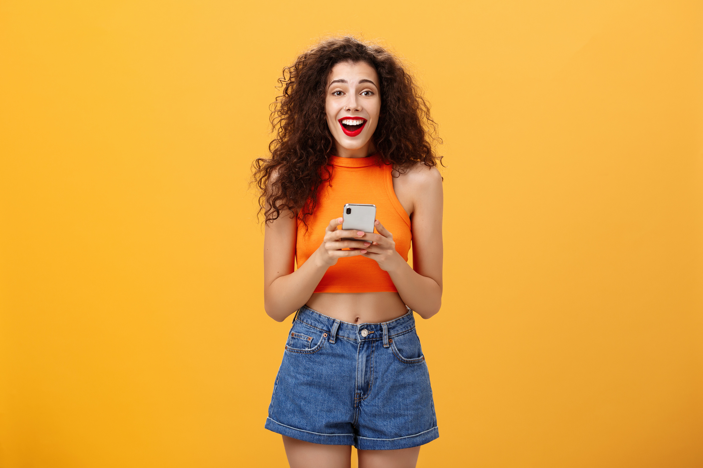 Indoor Shot of Surprised Happy and Delighted Attractive European Female with Curly Haircut in Cropped Top Holding Smartphone Gazing at Camera Amazed and Pleased Receiving Awesome News via Messages